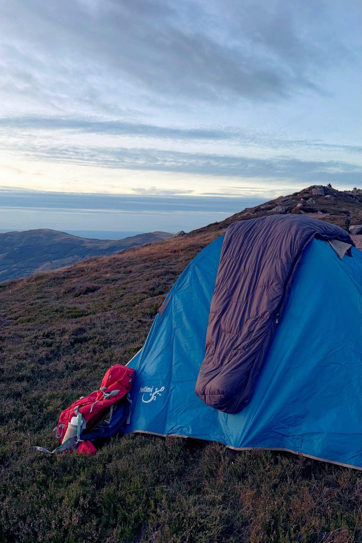 Tente 3 personnes Isis 3, marque Freetime, vue en fin de journée, sur un plateau de montagne, avec sac de couchage Lodenn posé dessus.
Crédit @cestmeline