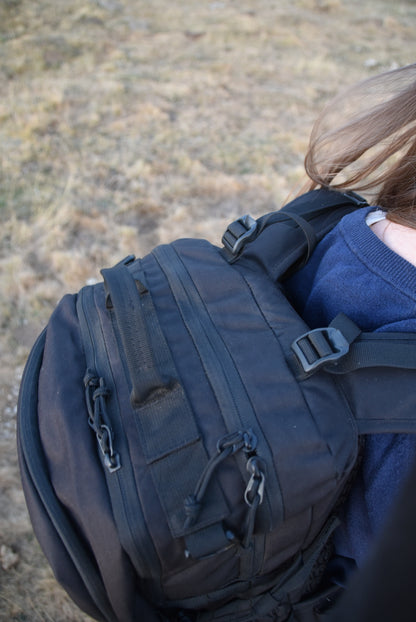 Sac d'évacuation Mangouste 32, marque Freetime, capacité 32 litres - sac noir avec système MOLLE et compartiment ordinateur, vu du dessus porté par une jeune femme - crédit @cestmeline